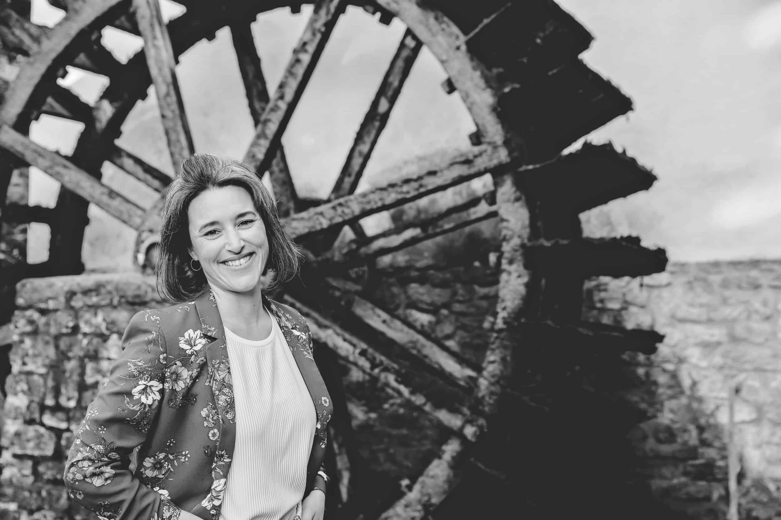 Schwarz-weiß-Foto einer lachenden Frau mit dunklem, schulterlangem Haar, die vor einer alten, verrosteten Wassermühle steht.