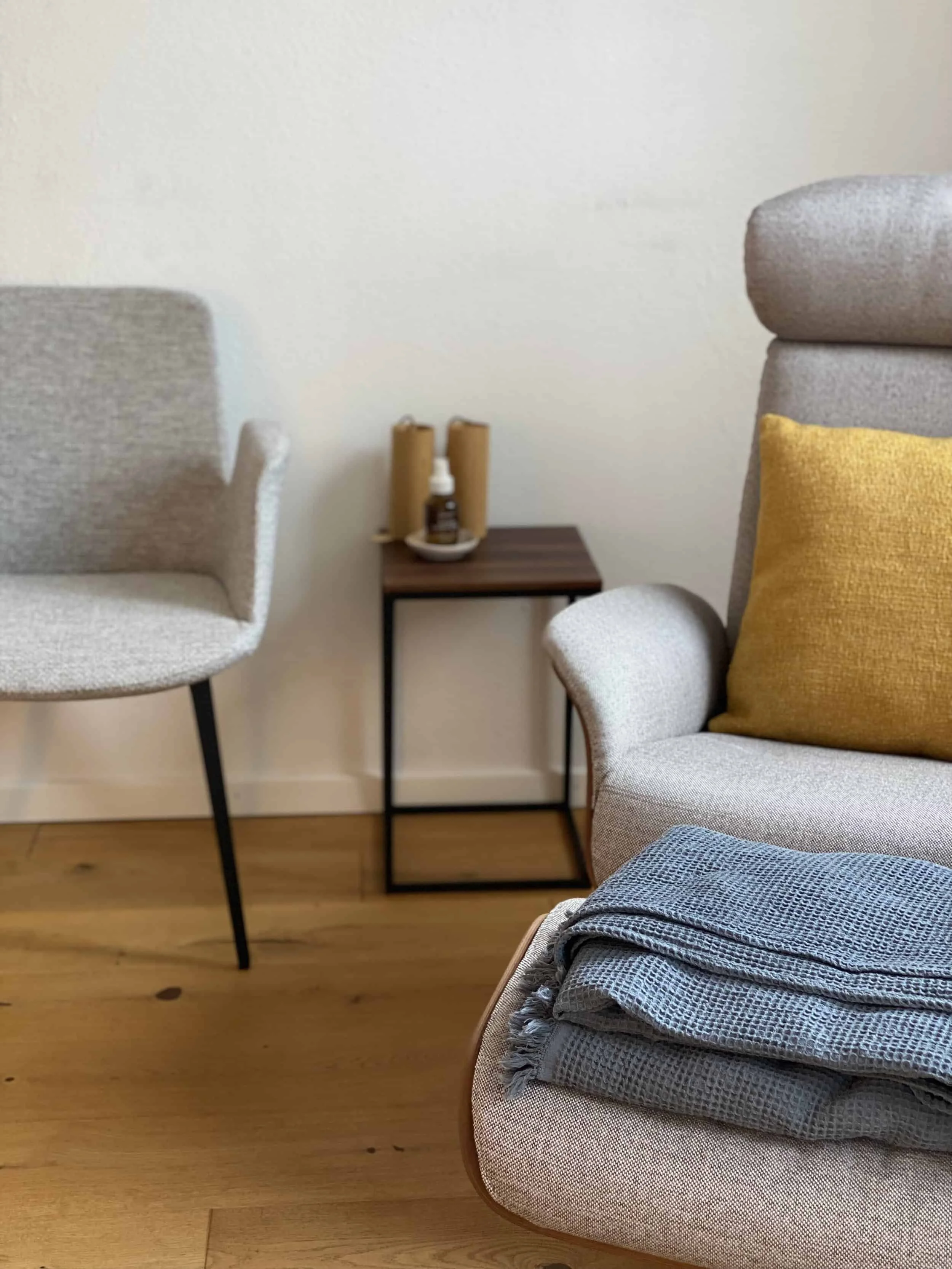 Gemütliches Wohnzimmer mit einem Beistelltisch, Stühlen, einem Sessel mit gelber Kissen und einem gefalteten grauen Decke auf einem Hocker