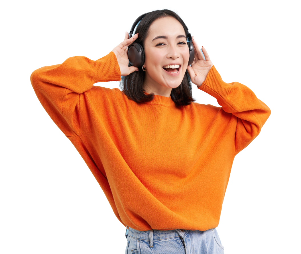 A young woman with dark hair, wearing an orange sweater and blue jeans, smiling happily while listening to music through large black headphones.