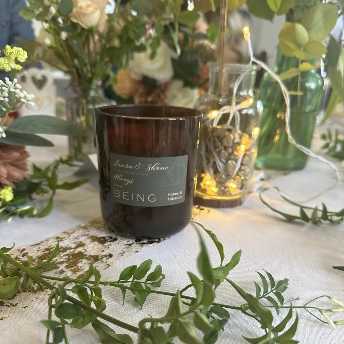 A dark glass candle labeled "Honey & Tobacco" on a table decorated with flowers, greenery, and a jar of string lights.