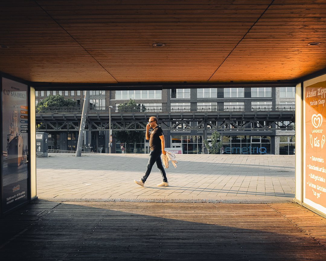 Ein Mann läuft auf einer Stadtstraße, während er ein Handy benutzt, durch eine unterirdische Passage mit Holzdecke. Auf beiden Seiten der Passage befinden sich Werbetafeln.