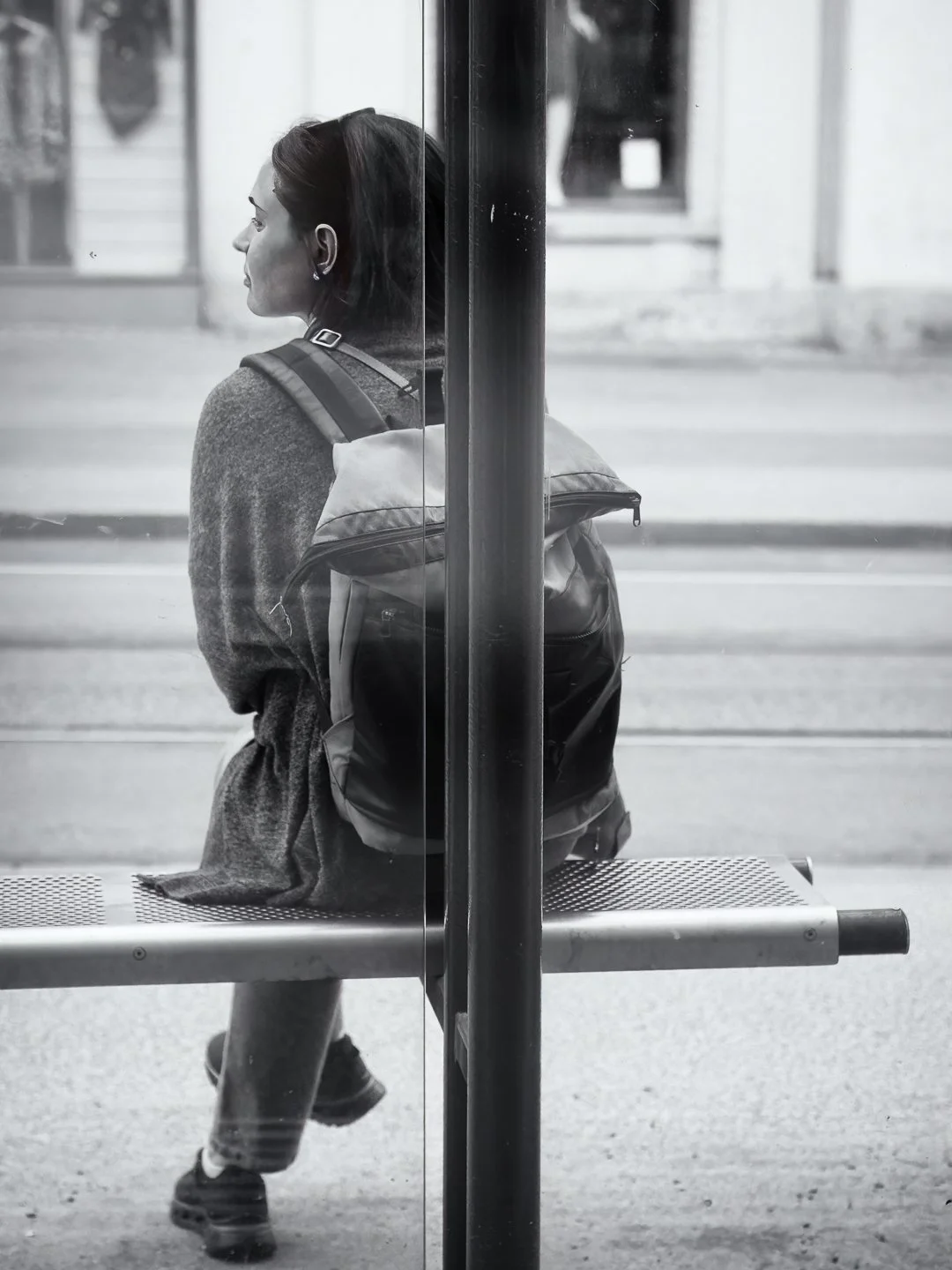 Frau sitzt an Bushaltestelle mit Rucksack, blickt nach links, schwarz-weiß Foto.