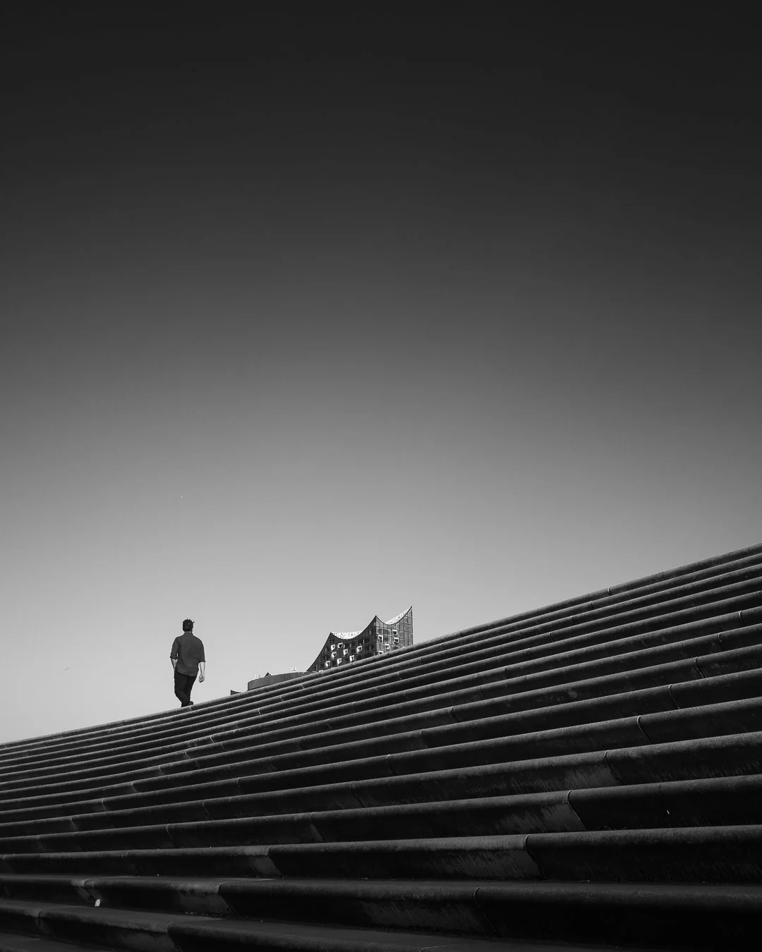 Ein Mann geht an einem steilen, gebogenen Treppenabsatz vorbei. Im Hintergrund ist ein modernes, unregelmäßig gestaltetes Gebäude sichtbar. Das Bild ist schwarz-weiß gehalten.