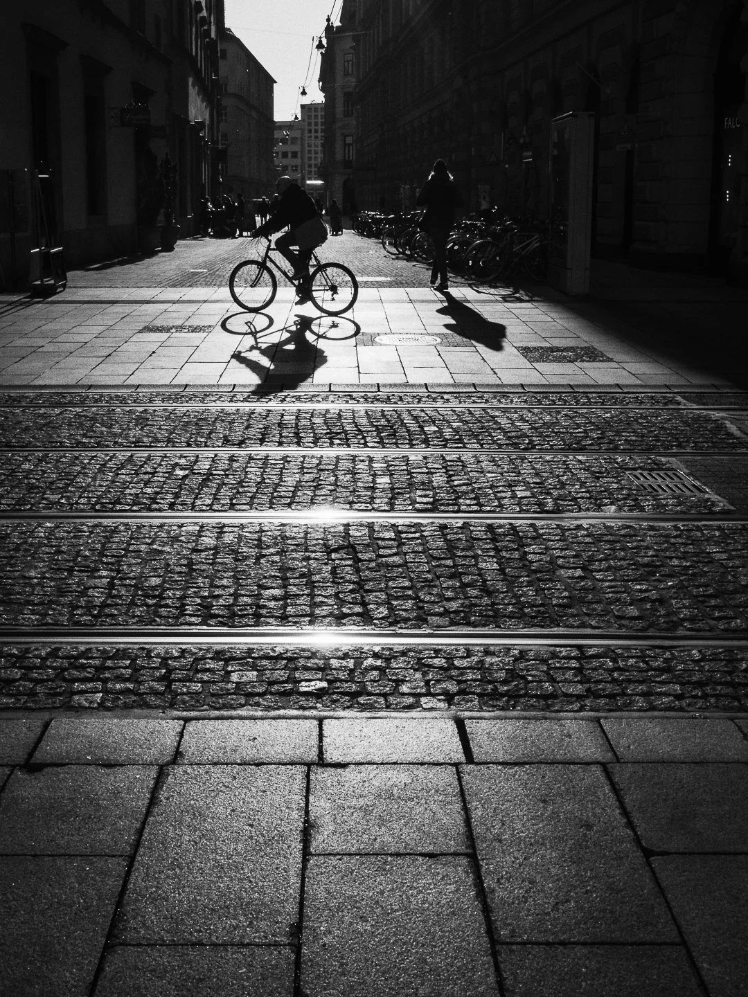 Schwarz-Weiß-Foto einer Stadtstraße im Sonnenlicht, mit einem Fahrradfahrer und einer Fußgängerin, die Schatten auf dem Pflaster und die Gebäude im Hintergrund sichtbar sind.