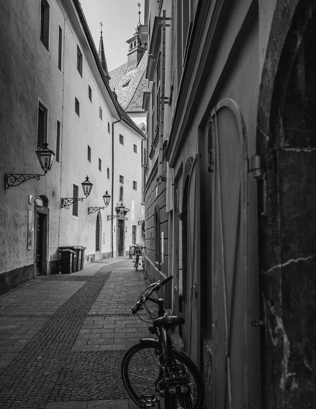 Ein schmaler, gepflasterter Straßenabschnitt in einer alten europäischen Stadt mit historischen Gebäuden, Laternen und Fahrrädern in schwarz-weiß.