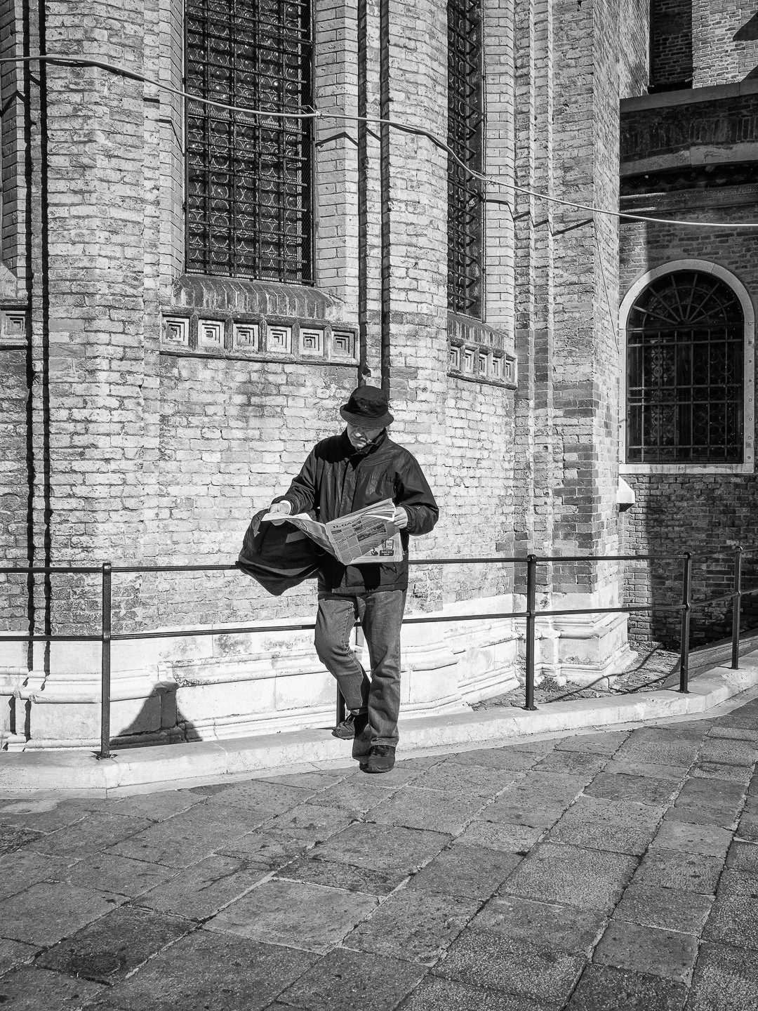 Ein Mann liest eine Zeitung, lehnt an einer alten Ziegelmauer mit großen fenstern, in Schwarz-Weiß-Fotografie.