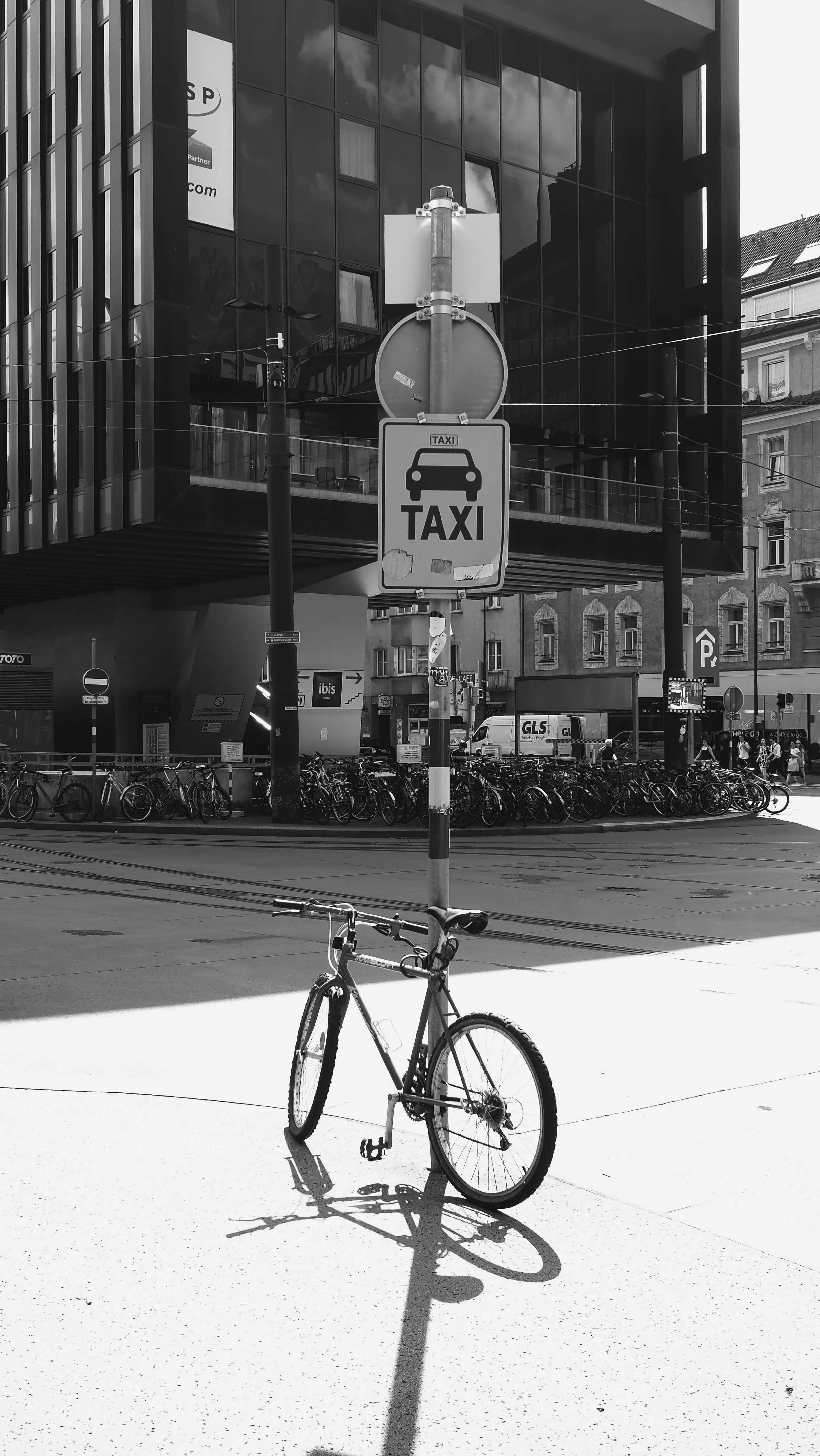 Ein Fahrrad steht an einem Laternenpfahl mit Taxi-Schild in einer Stadt. Im Hintergrund sind weitere Fahrräder, Fahrzeuge und moderne Gebäude.