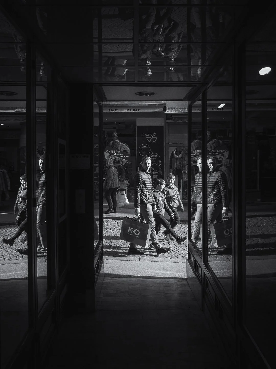 Schwarz-Weiß-Foto von Menschen, die an einem Laden vorbeigehen, vom Eingang eines Gebäudes aus gesehen, mit reflektierenden Glasspeigeln