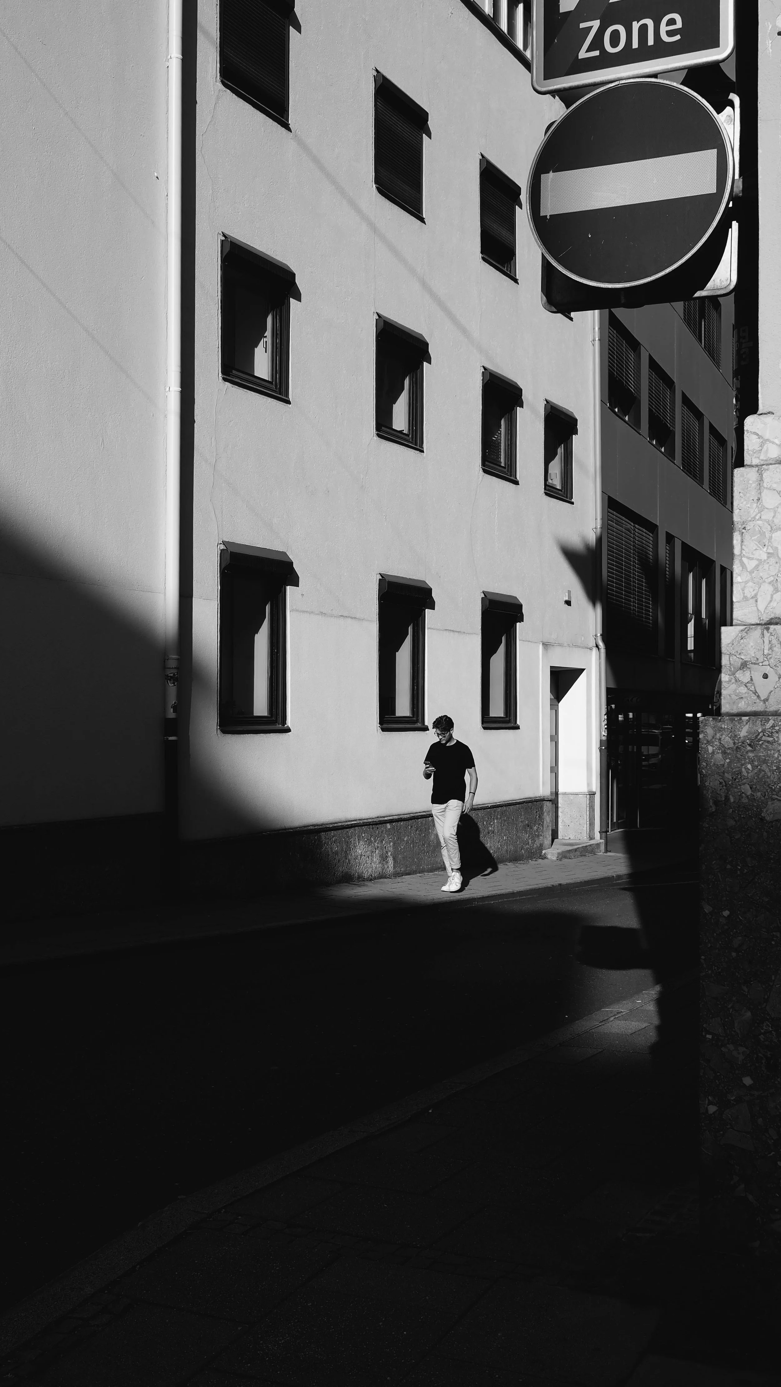 Ein junger Mann in Schwarzen T-Shirt und hellen Hosen läuft auf einem schattigen Stadtweg, während er auf sein Smartphone schaut, neben einem mehrstöckigen Gebäude mit mehreren Fenstern, geprägt von Kontrasten zwischen Sonnenlicht und Schatten.