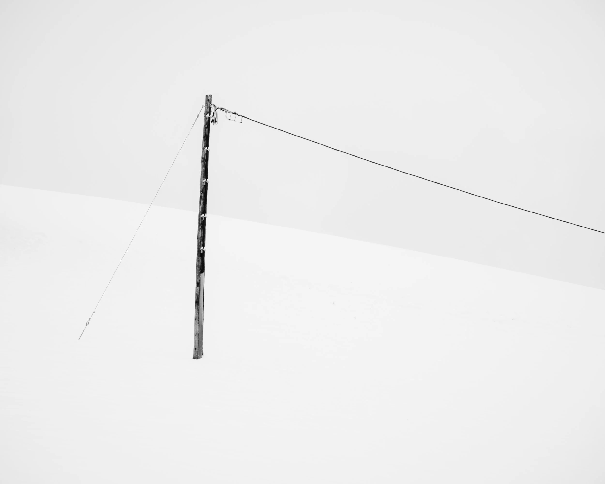 Ein einzelner Holzpfosten steht auf schneebedegnem Gelände, mit einer fixierten Laterne oben. Die Umgebung ist winterlich, und der Himmel ist bewölkt.