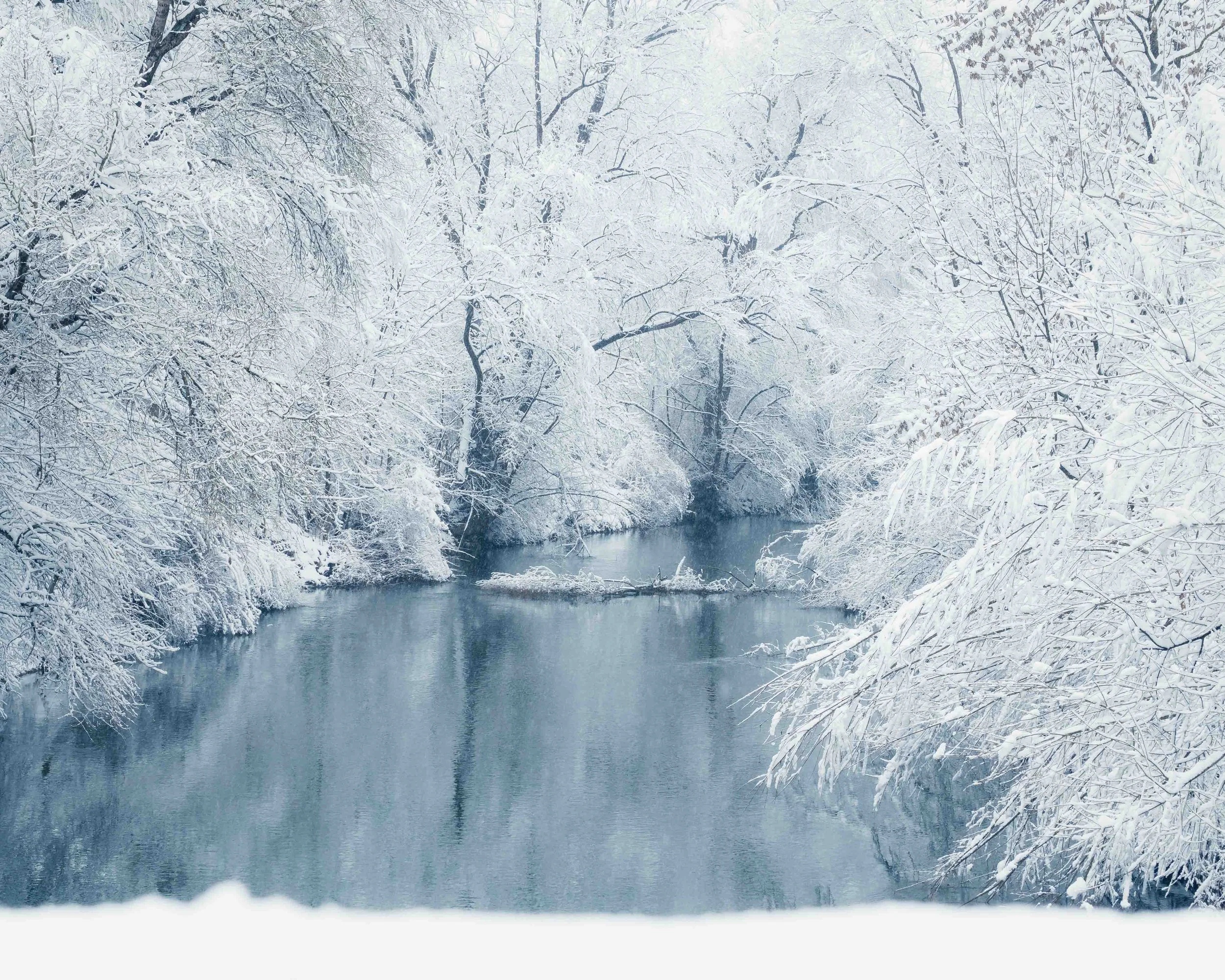 Ein verschneiter Fluss, umgeben von Bäumen mit Schneebedeigten Ästen in einer Winterlandschaft