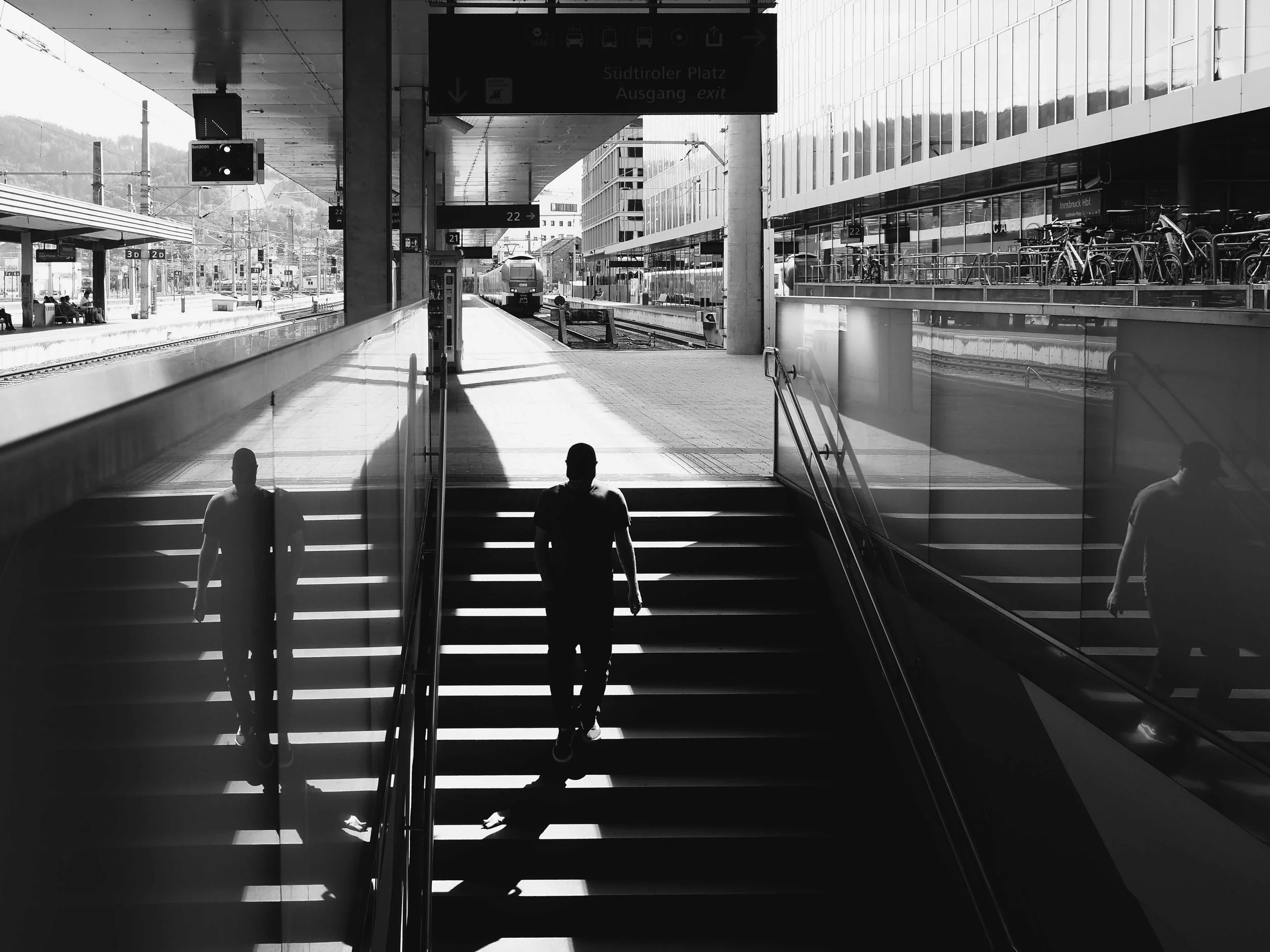 Schwarz-Weiß-Foto eines Bahnhofs mit zwei Männern, die die Treppe hinaufgehen, reflektiert im Glas. Im Hintergrund sind Züge, Fahrradständer und ein Gebäude sichtbar.