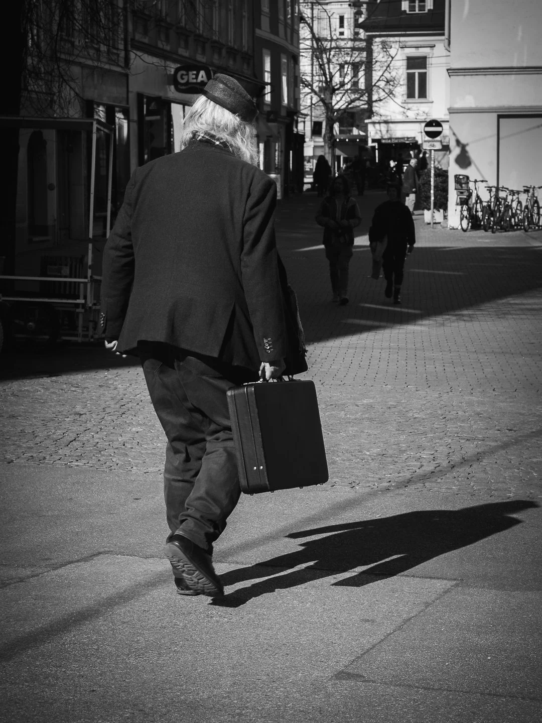 Schwarz-Weiß-Fotografie eines älteren Mannes mit grauem Haar, der einen schwarzen Koffer in der Hand hält und die Straße überquert, mit mehreren Fußgängern im Hintergrund.