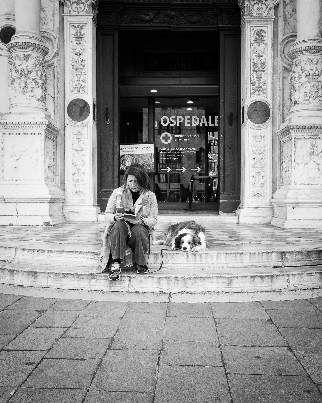Eine Frau sitzt vor einem Krankenhaus, liest ein Buch und hat einen Hund neben sich liegend. Das Krankenhaus hat eine barocke Fassade mit dekorativen Säulen und Verzierungen. Die Tür zeigt die Aufschrift 'Ospedale'.