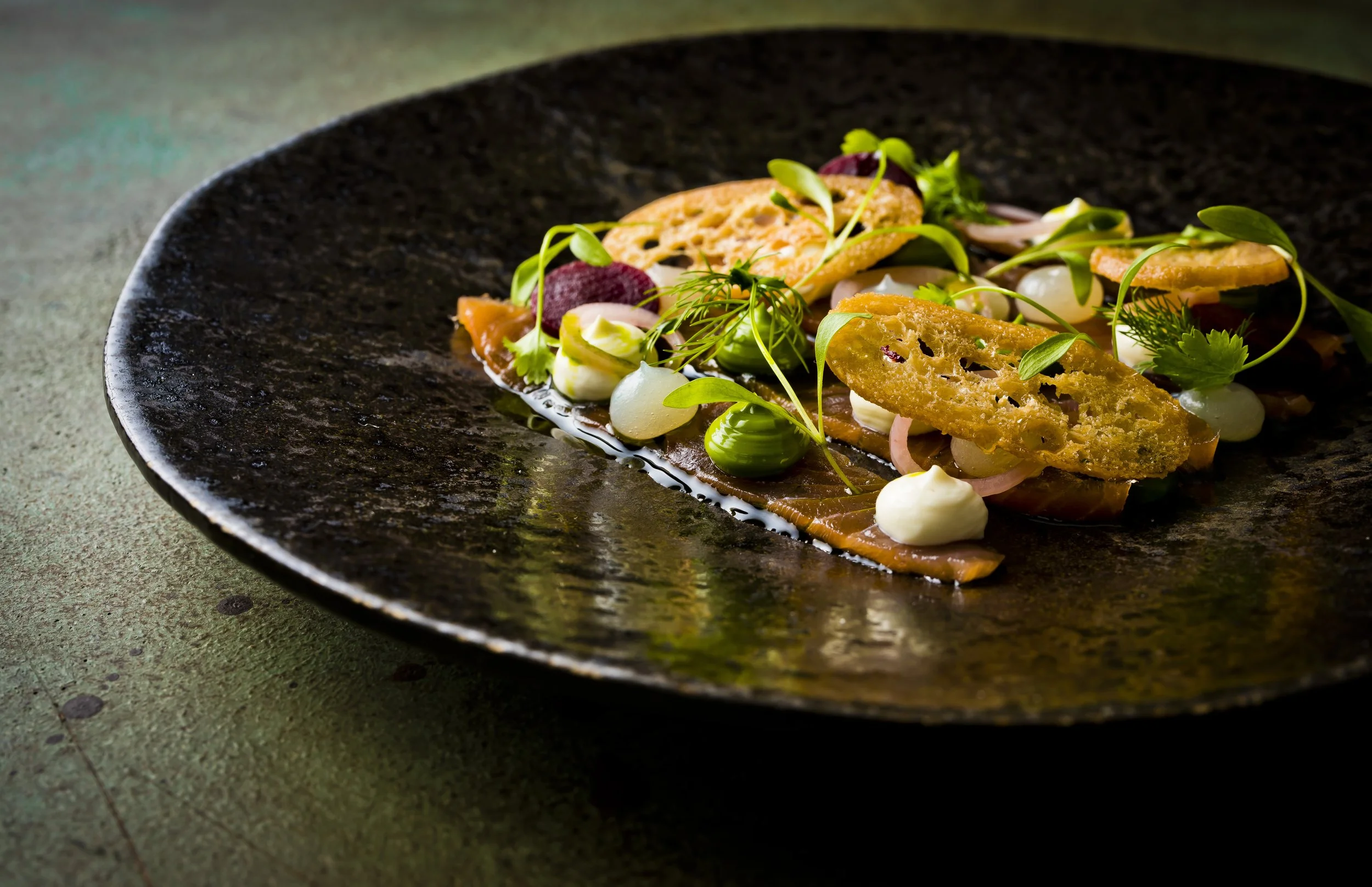 Gourmet dish with smoked salmon, crispy bread, creamy dollops, greens on dark plate