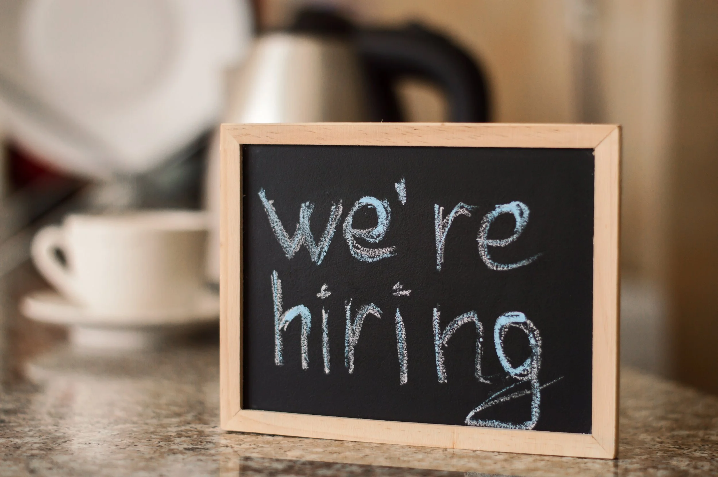 Chalkboard sign with "we're hiring" written on it, placed on a kitchen counter next to a teacup and electric kettle.