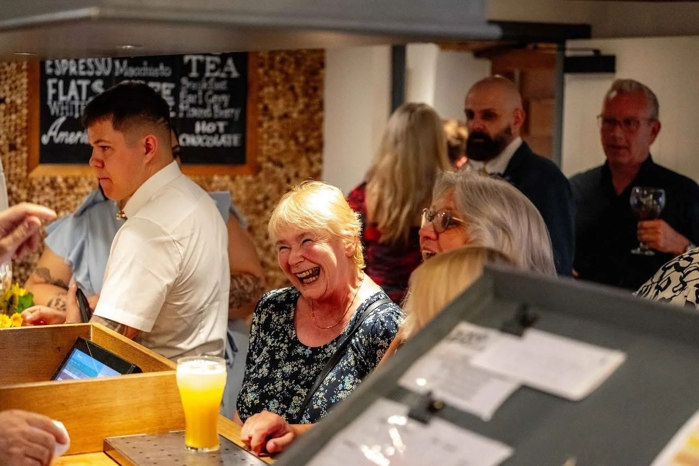 People socializing at a bar, with a woman laughing and others engaged in conversation. A bar counter with a glass of beer is visible.