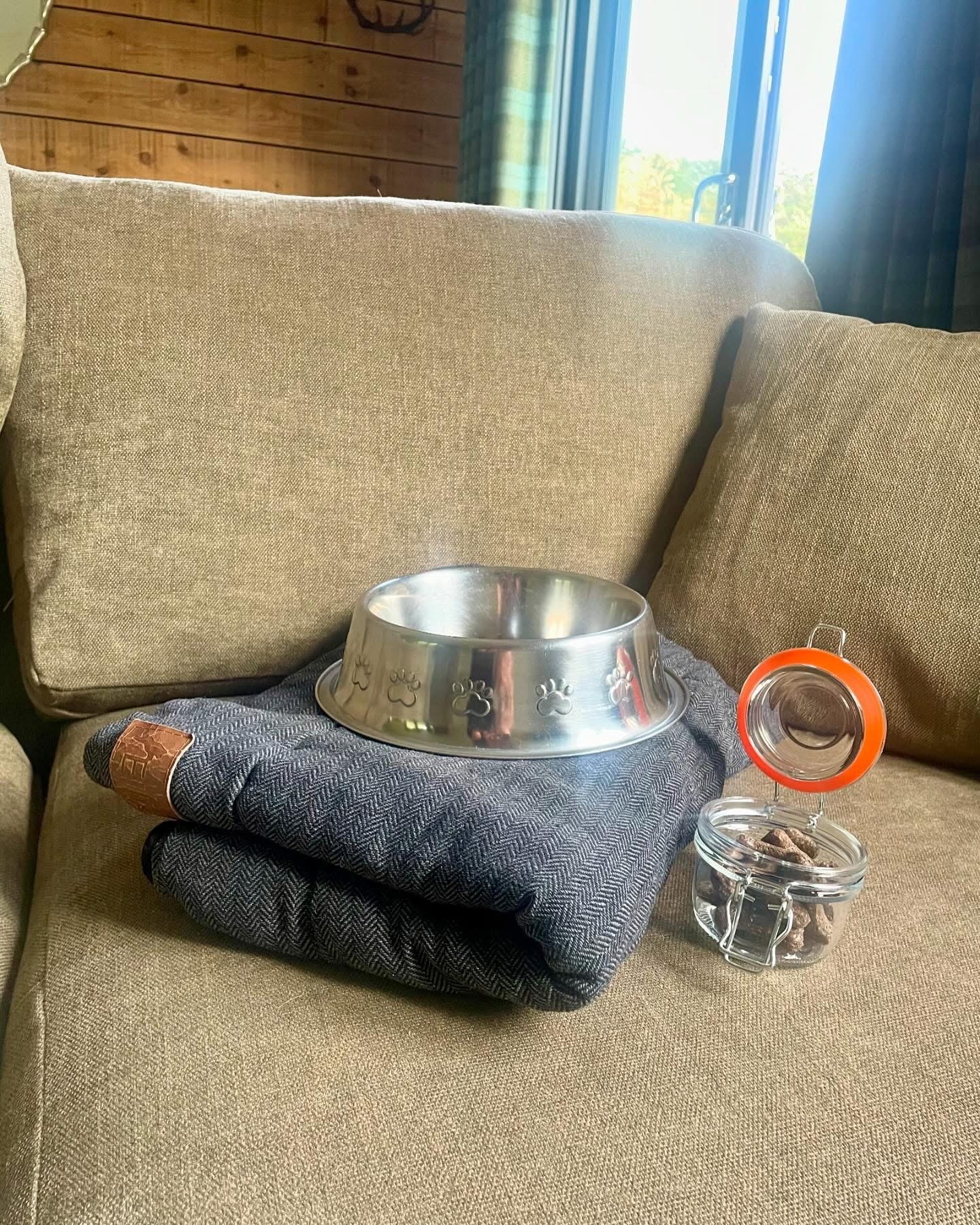 A pet setup on a couch, featuring a gray folded blanket, a metal dog bowl with paw prints, and a small glass jar of dog treats.