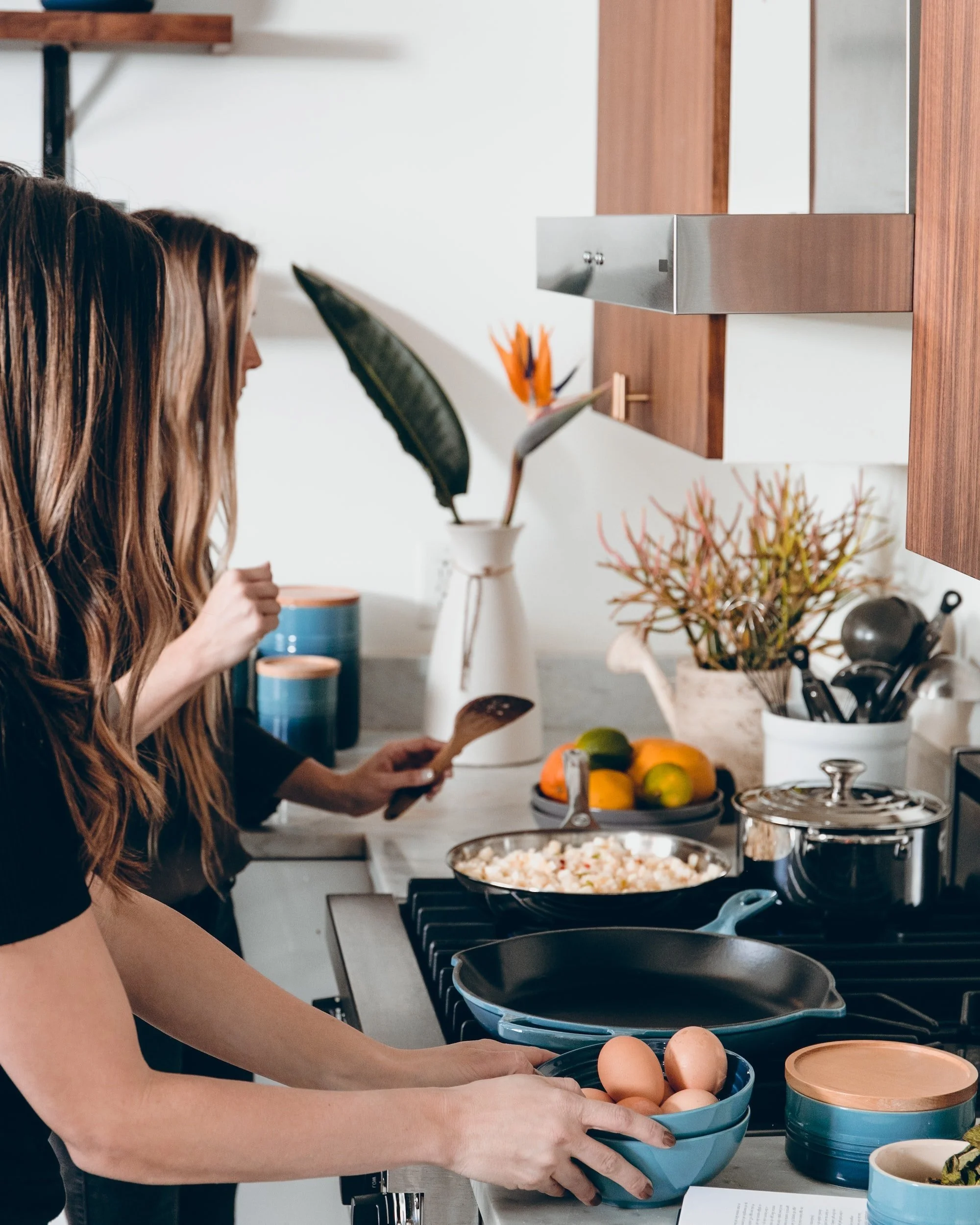 Two women cooking in a kitchen with eggs, fruits, and a skillet on the stove.