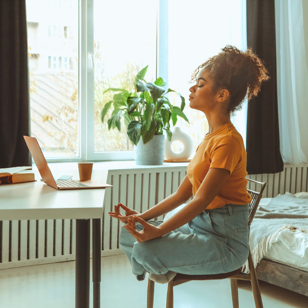 Woman practicing meditation in her bedroom near a window with sunlight, sitting cross-legged on a chair, with a laptop, books, plant, and decorative vase nearby.