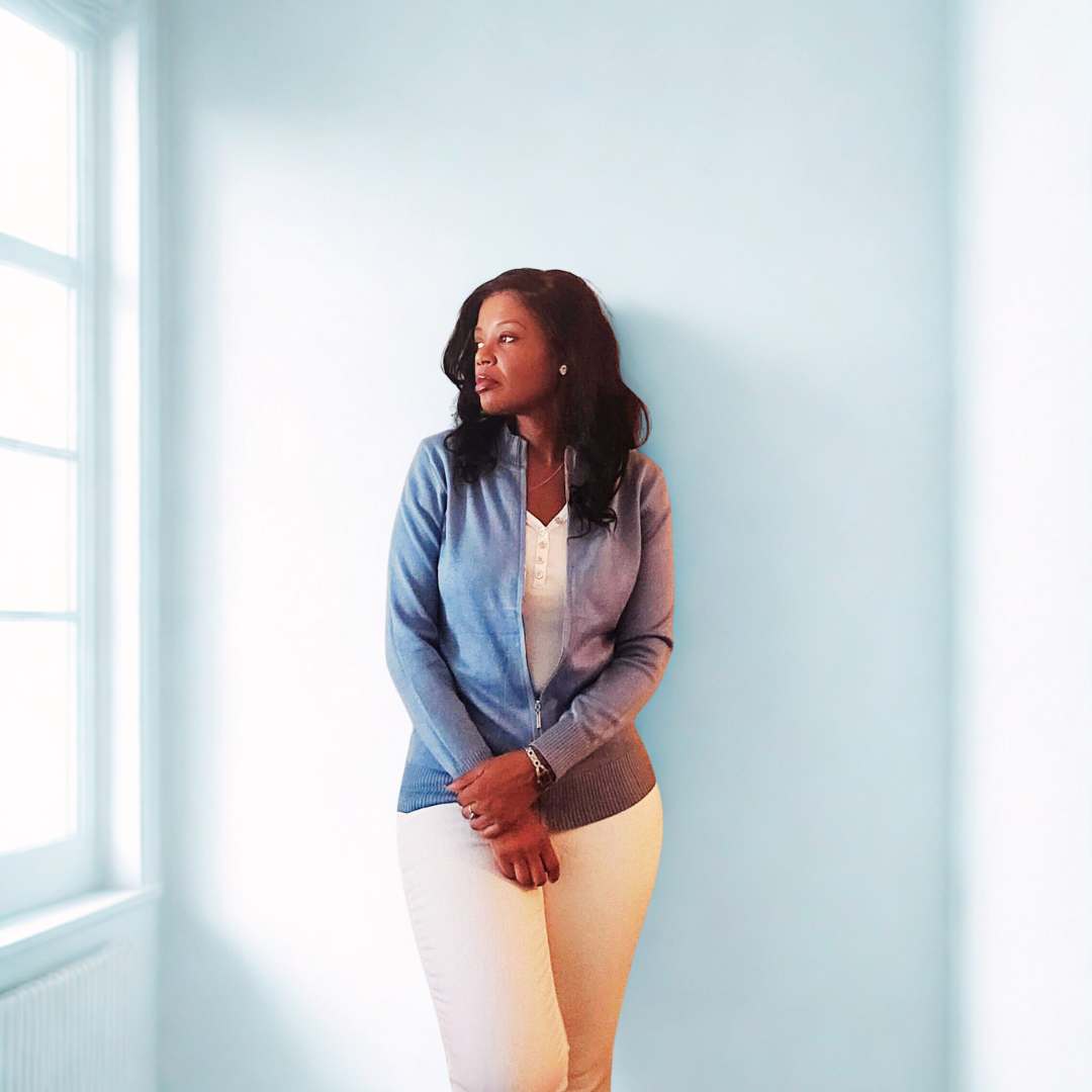 A woman standing in a bright room with light blue walls and a large window, looking to her left, wearing a gray sweater over a white top and cream-colored pants.