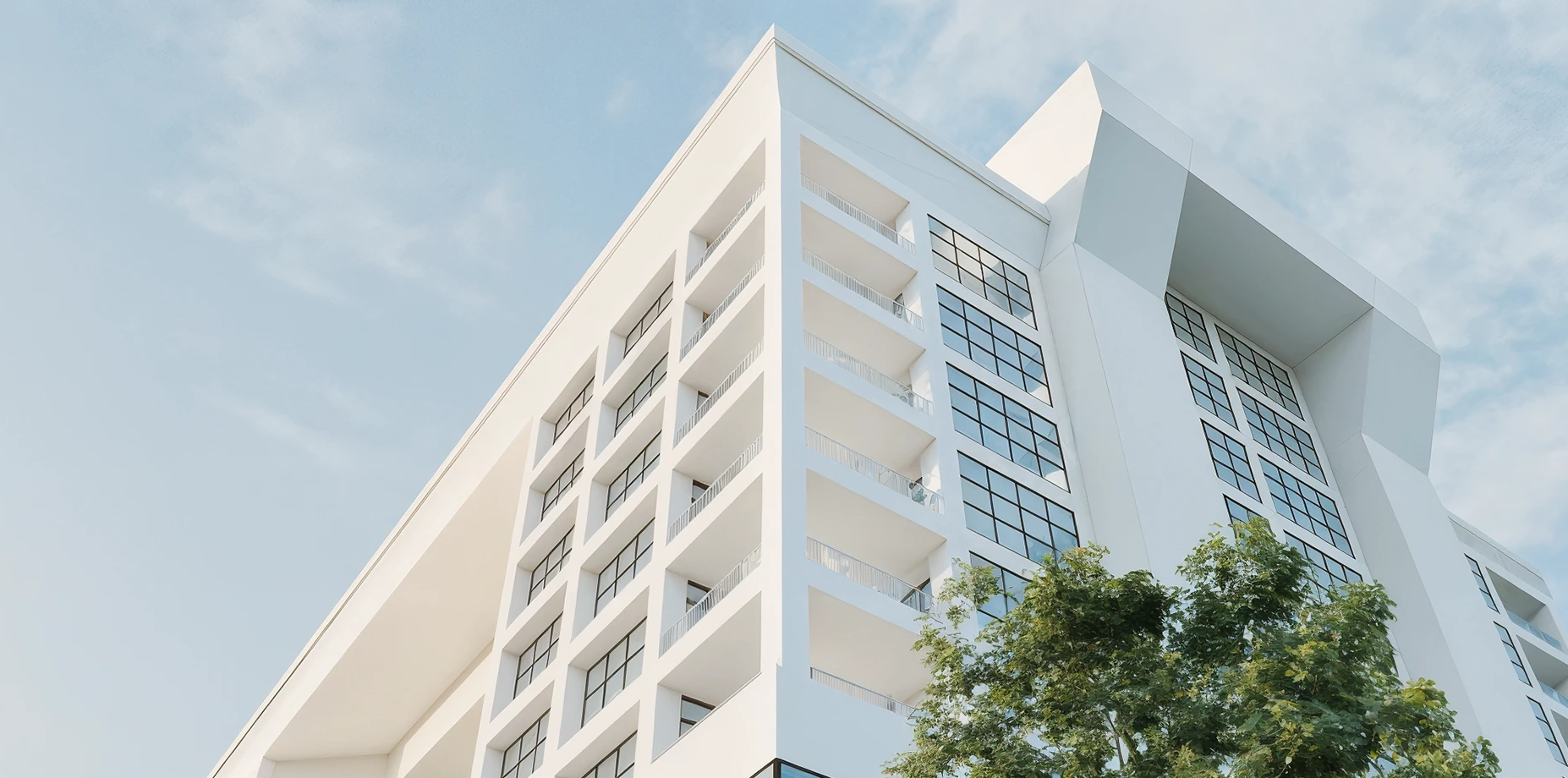 Modern white multi-story building with many large windows and balconies, against a blue sky with some clouds, with a green tree in the foreground.
