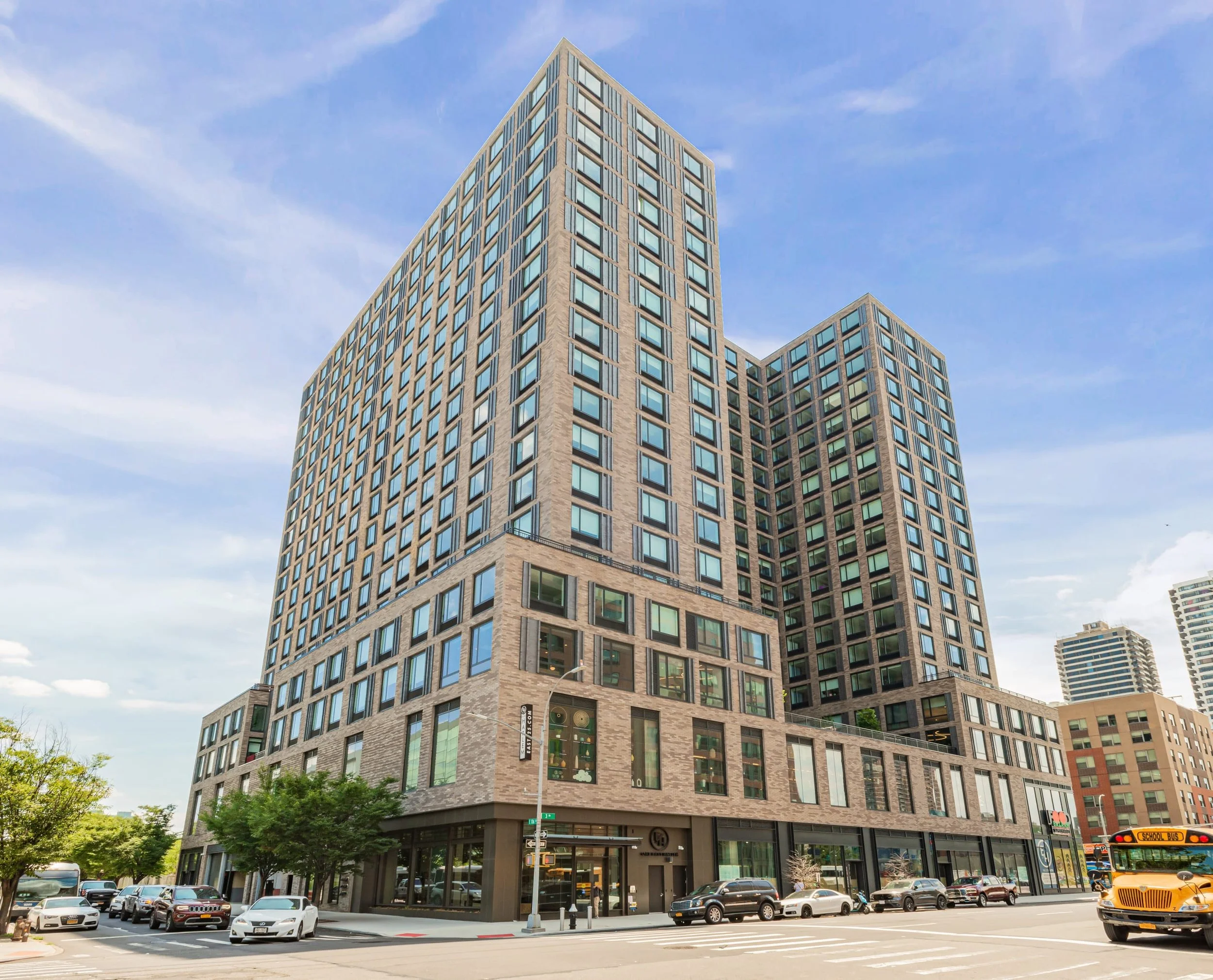 A modern multi-story commercial building with large glass windows and a brick exterior, situated at a city street corner with cars parked and a school bus in the background.