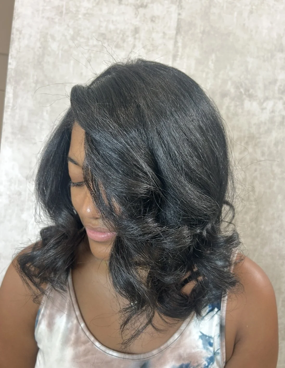 A woman with shoulder-length wavy black hair is looking down and to her left, wearing a sleeveless top with a tie-dye pattern.