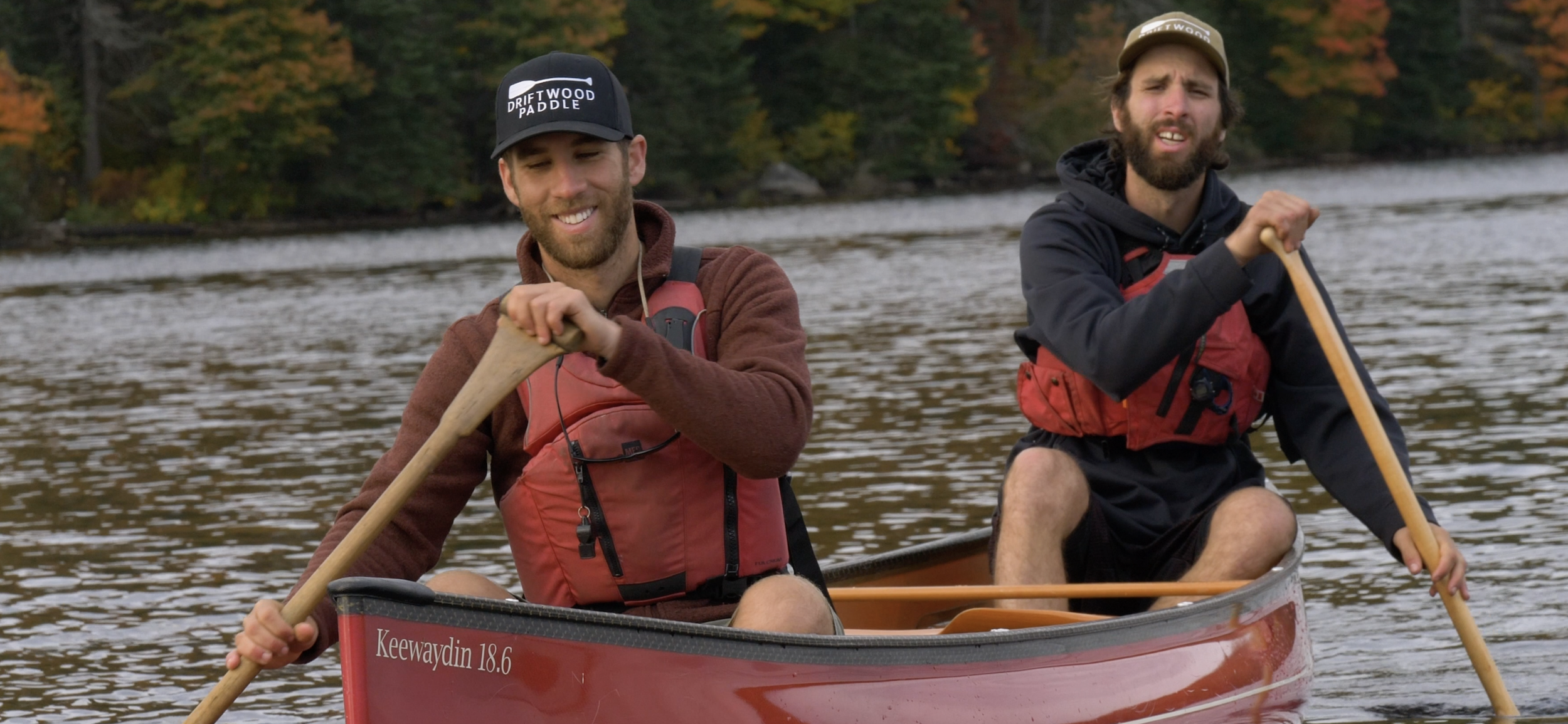 Two men paddling a red canoe on a river, surrounded by fall foliage, both smiling and wearing life jackets and casual clothing.