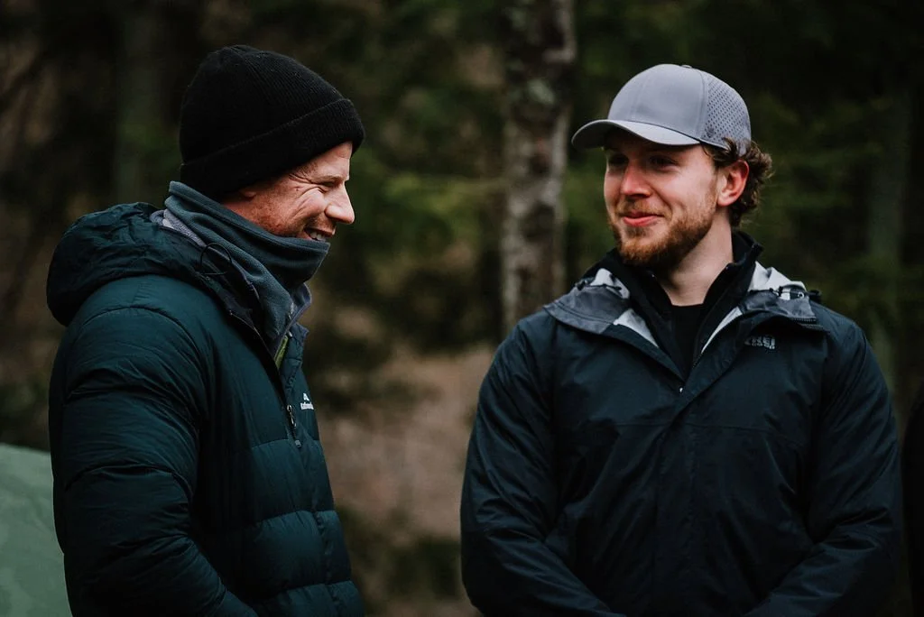 Two men outdoors in a forest, smiling and talking, dressed in hiking jackets and hats.