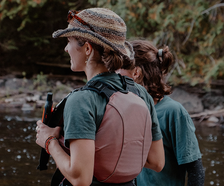 Two women hiking by a river, one wearing a hat and carrying a backpack, the other with curly hair in a ponytail.