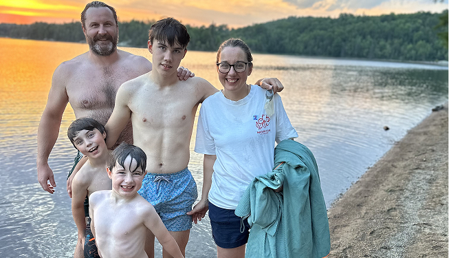 Family of five by a lake during sunset, smiling and posing together in swimwear and casual clothes.