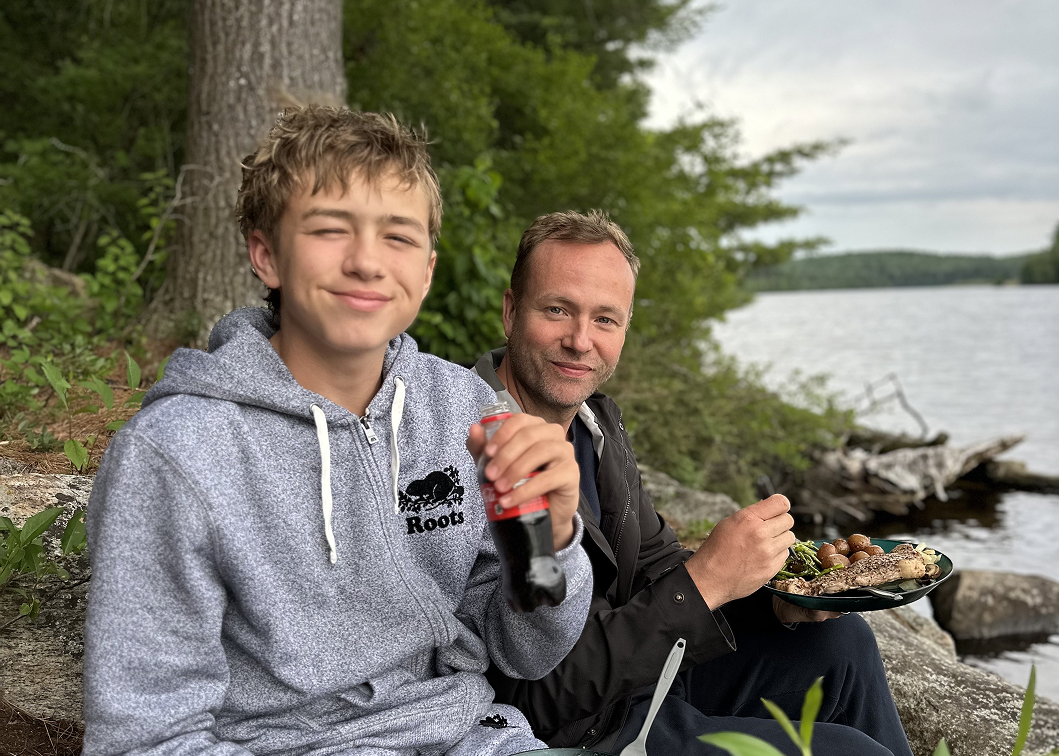 A young boy and an older man sitting outdoors by a lake, enjoying a meal and drinks. The boy is smiling and holding a soda, while the man is eating food from a plate. They are surrounded by trees and rocks, with a cloudy sky overhead.