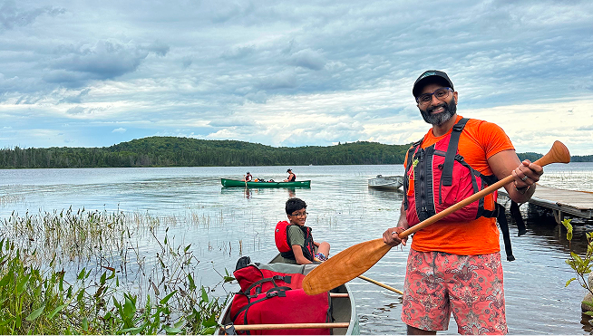 A man and two children prepare for a canoe trip on a lake with a wooded shoreline, with cloudy skies overhead.