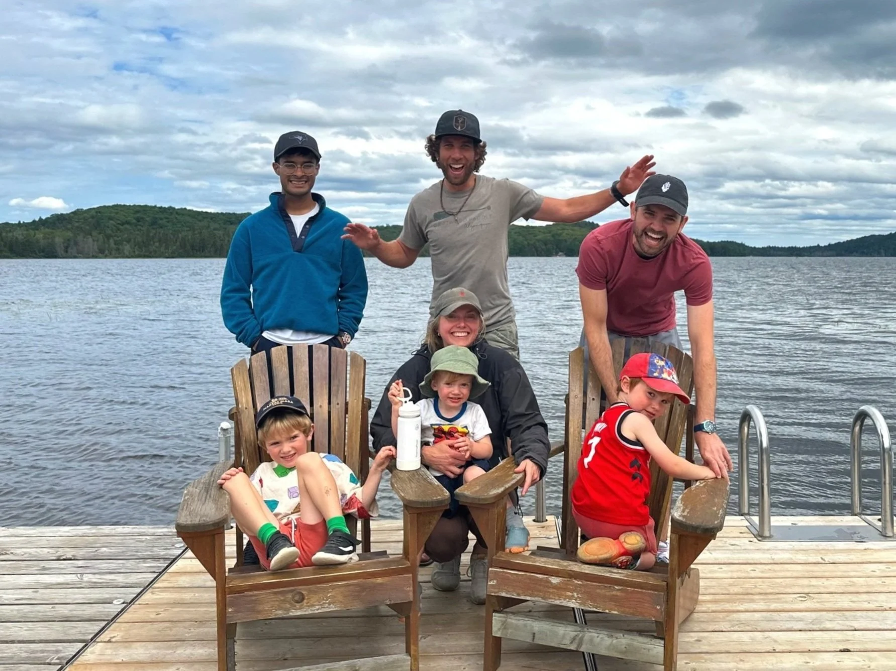 Group of five smiling people, including three children, sitting and standing on a wooden dock by a lake with a tree-covered shoreline, during daytime with cloudy sky.