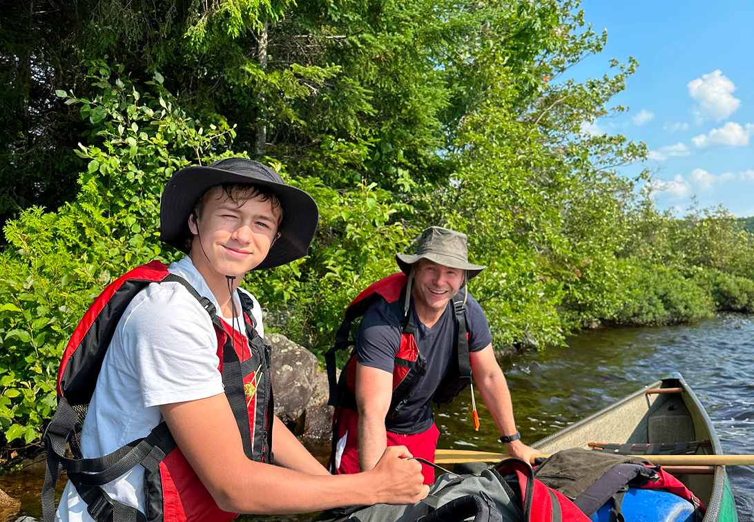 Two smiling males wearing hats and life jackets on a canoe near the water's edge, surrounded by green trees and blue sky.