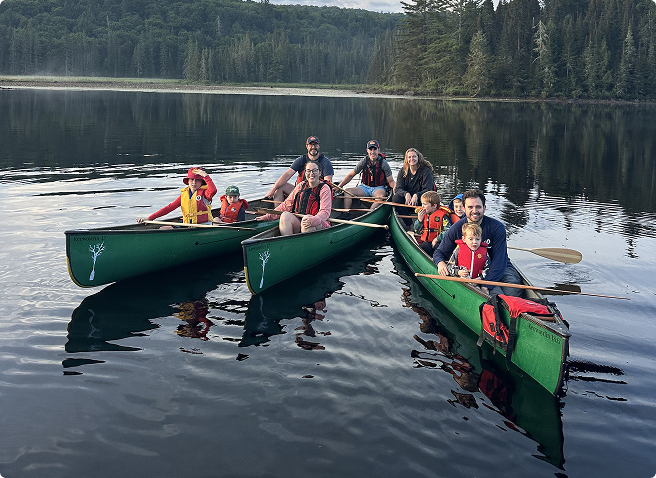Group of people, including children and adults, enjoying a canoe trip on a calm lake surrounded by trees.