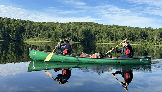 Two people in a green canoe paddling on a calm lake with a forested shoreline under a partly cloudy sky.