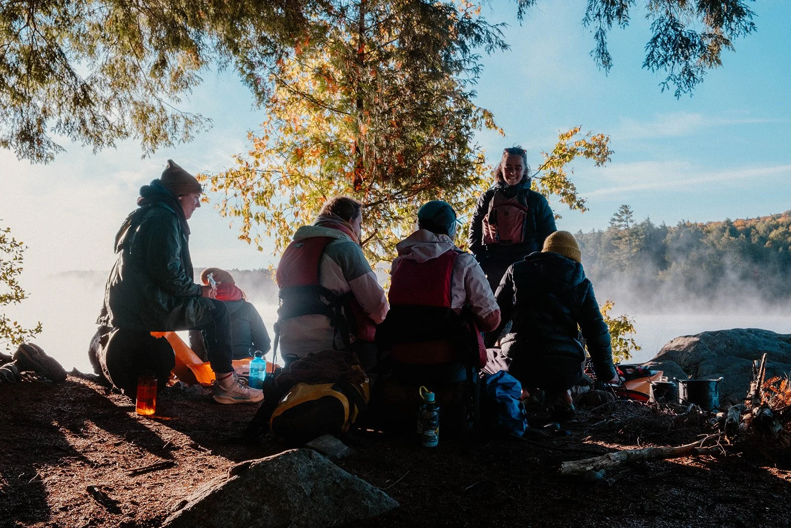 A group of people on a camping trip by a lake during sunrise, sitting on the ground with backpacks and camping gear, under a tree with mist rising from the water in the background.