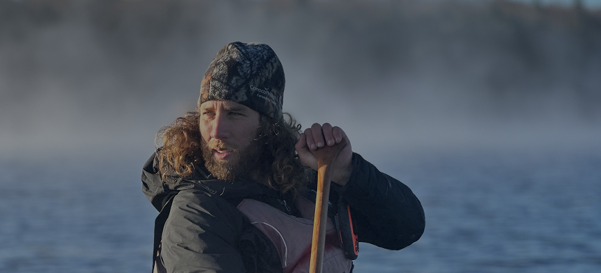 Man with long curly hair and beard wearing a camouflage beanie and jacket, paddling a canoe or kayak on a misty lake in the early morning