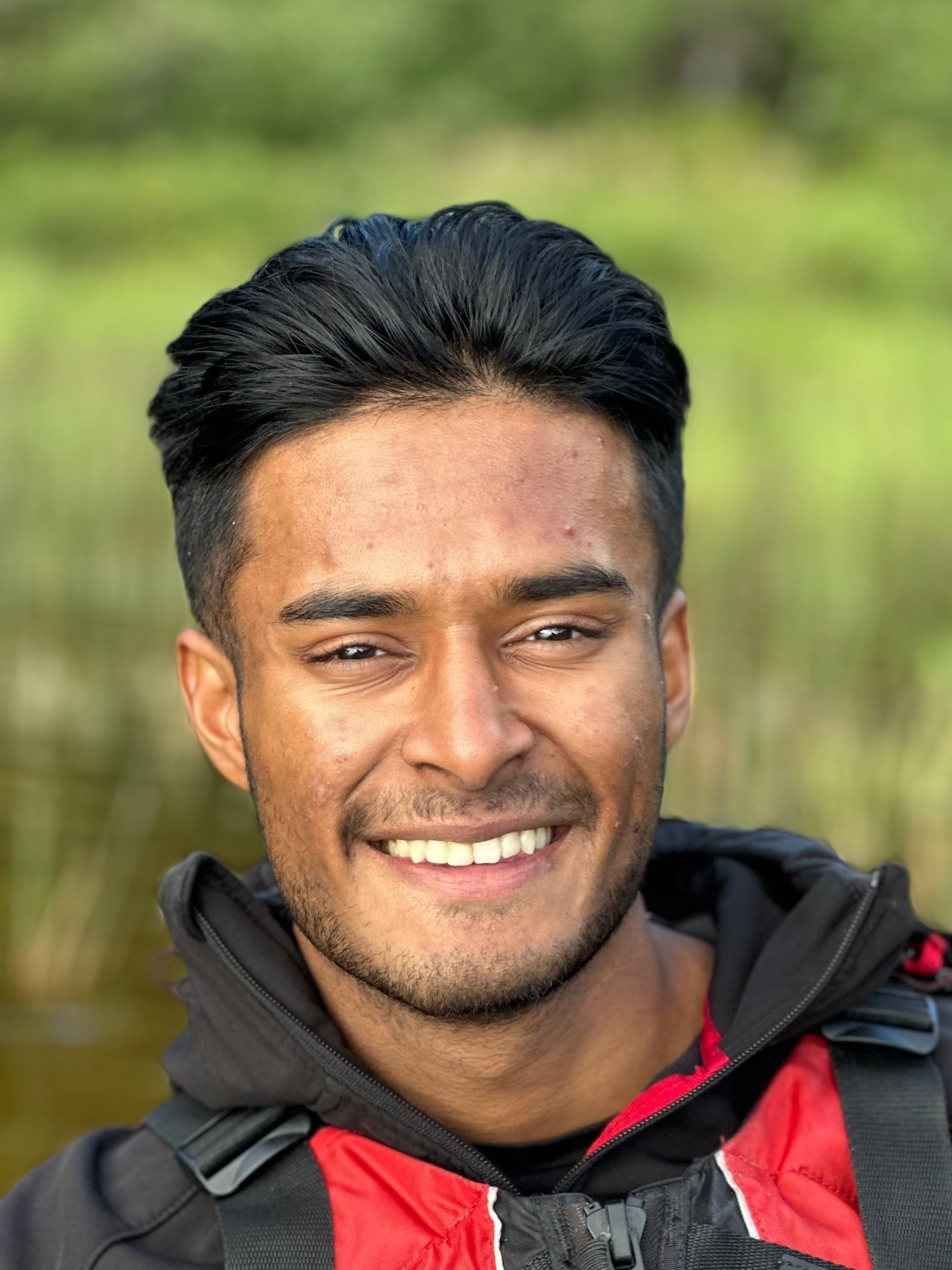 Close-up of a smiling young man with short black hair, light brown skin, wearing a black and red jacket, outdoors with green foliage in the background.
