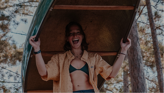A woman smiling and holding up a canoe lid, standing outdoors among trees.