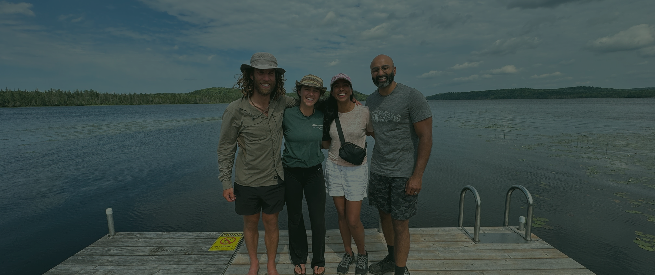 Four people standing together on a dock by a lake, smiling, with water and trees in the background on a partly cloudy day.