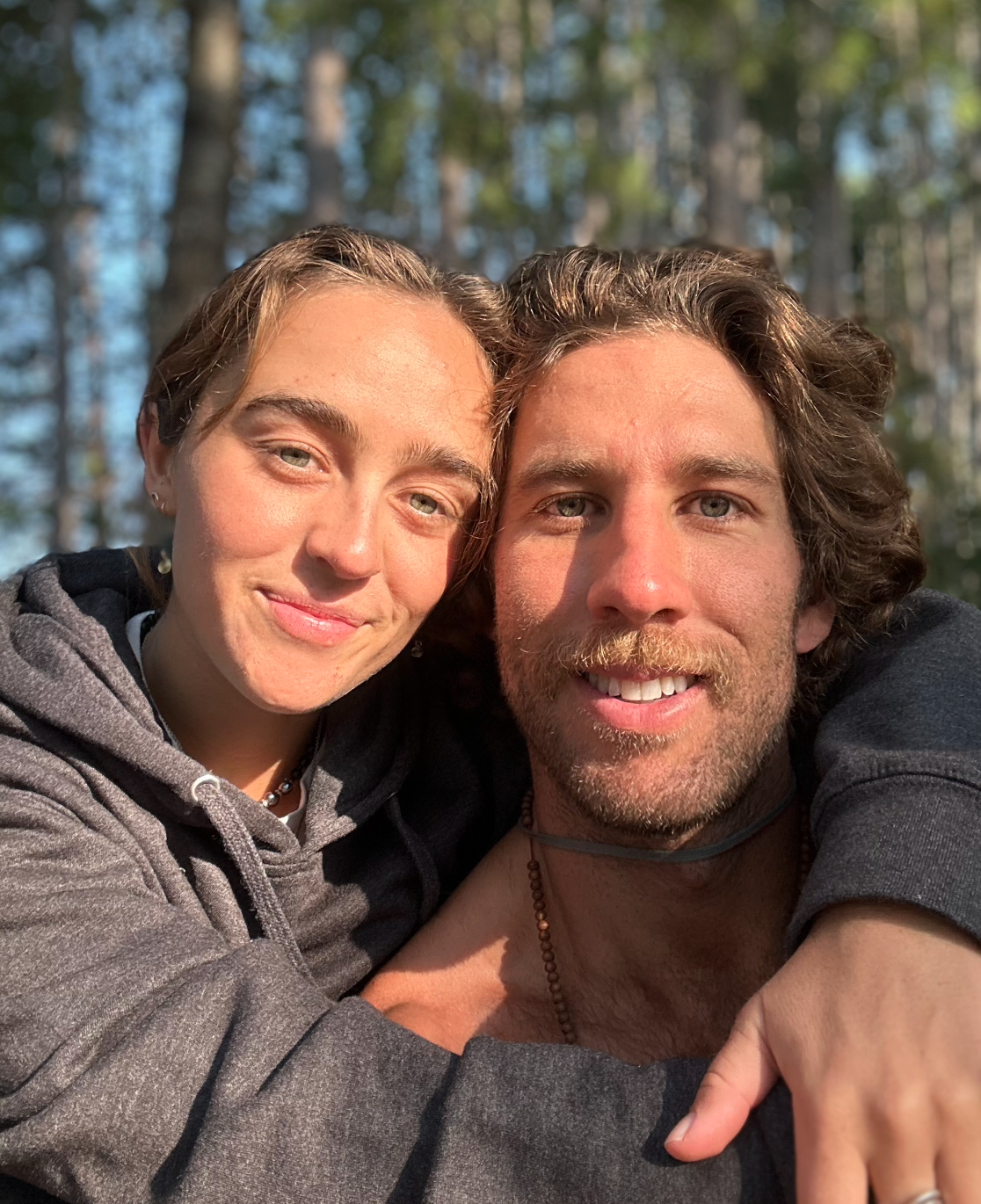 A young woman and a man smiling closely together outdoors in a wooded area with sunlight.