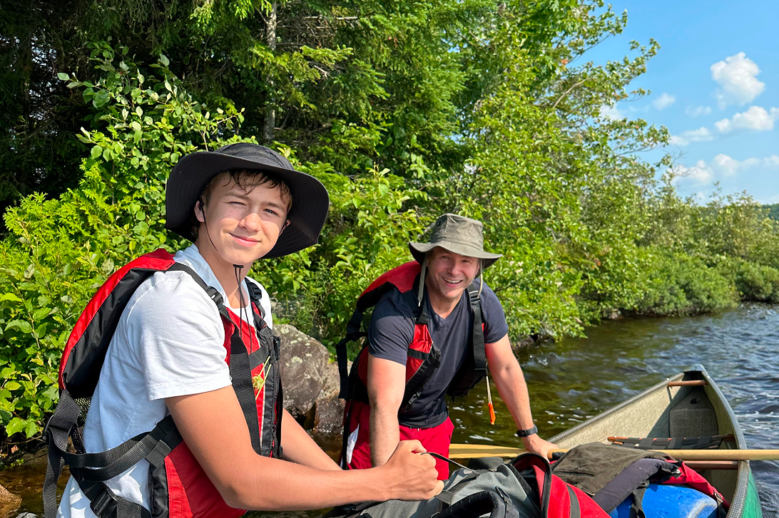 Two men preparing to canoe on a lake surrounded by lush green trees, wearing hats and life jackets.