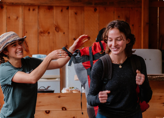 Two young women smiling and greeting each other inside a rustic wooden cabin, with backpacks, indicating a camping or outdoor trip.