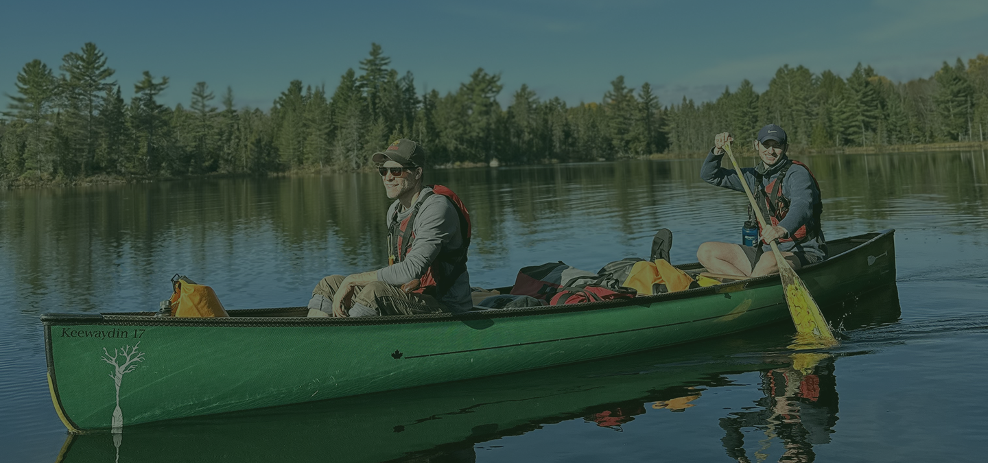 Two men in a green canoe paddling on a calm lake with a forest of pine trees in the background, smiling and enjoying a sunny day.