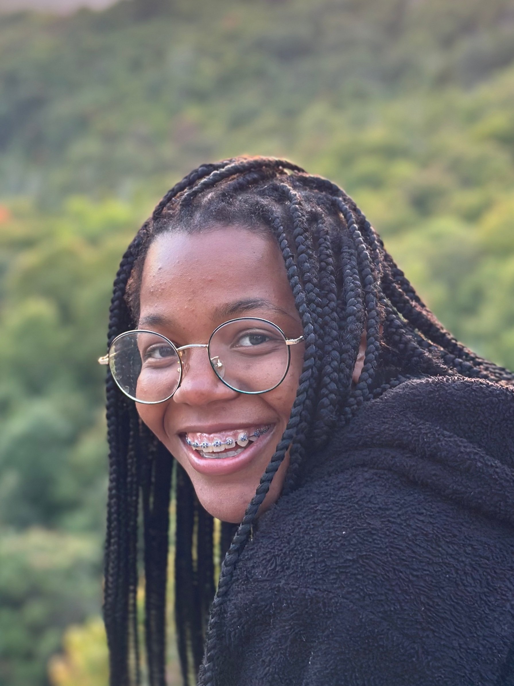 A young woman with long braided hair, glasses, and braces smiling outdoors with a green, blurred natural background.