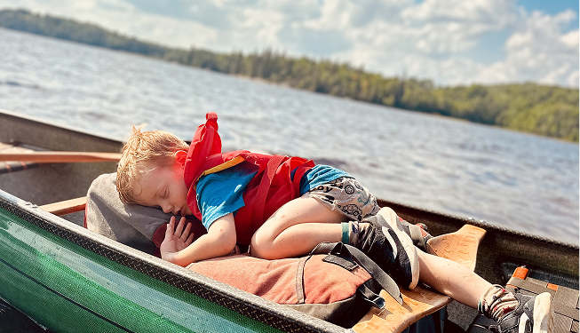 A young boy sleeping on a boat with a lake and trees in the background.