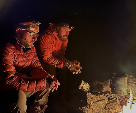 Two men wearing headlamps and outdoor gear sitting near a campfire at night.